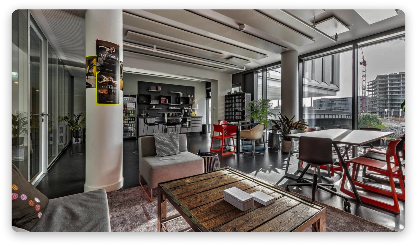 Foto eines offenen Büros mit hohen Decken und großen Fenstern, die einen Blick auf die Stadt bieten. Der Raum umfasst eine Lounge-Ecke mit einem Tisch, einer Kaffeebar, Möbel im Industrie-Stil, einer Kaffeemaschine und Pflanzen. Die moderne Architektur zeichnet sich durch einen Betonboden und eine Beton-Decke aus.